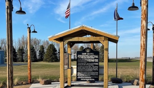 A wooden structure with a plaque about Camp Algona, surrounded by flags and green grass under a blue sky.