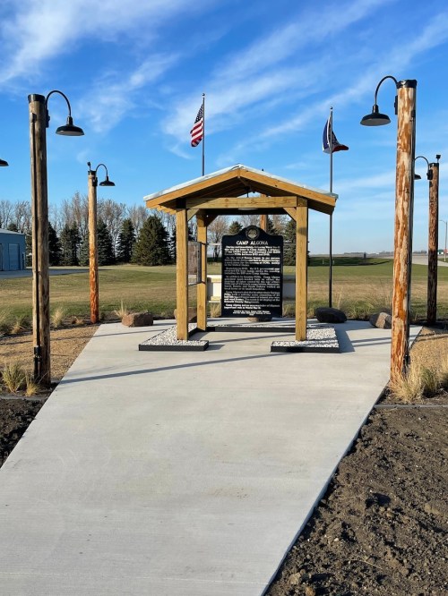 A memorial structure with a black plaque, surrounded by flags and lamp posts, set against a clear blue sky.