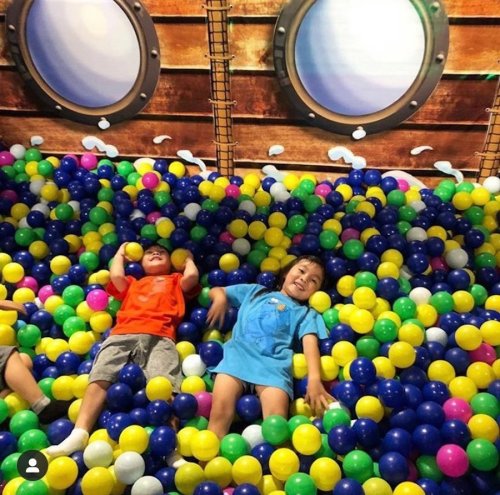 Two children play joyfully in a colorful ball pit filled with blue, yellow, and green balls, smiling and having fun.