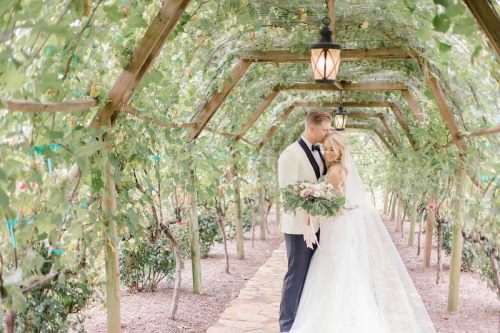 A bride and groom embrace under a lush, green arbor, surrounded by flowers and soft lantern light.