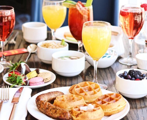 A vibrant brunch spread featuring waffles, sausage, fruit, and colorful drinks on a wooden table.