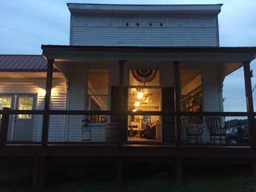 A cozy, illuminated porch of a white wooden building at dusk, featuring a welcoming entrance and rustic decor.