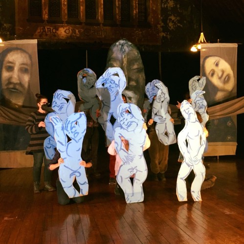 A group of people in costumes with blue silhouettes, performing in a dimly lit theater with large backdrop images.