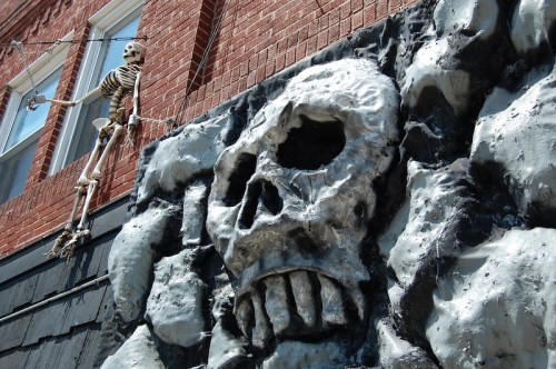 A large, silver skull sculpture on a brick wall, with a skeleton hanging nearby.