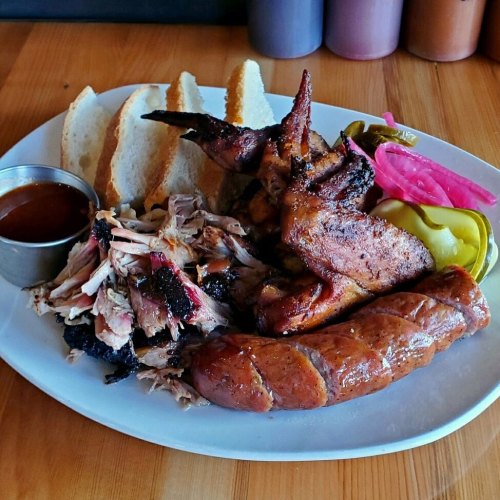 A plate of barbecue featuring smoked chicken wings, pulled pork, sausage, pickles, and slices of bread with sauce.