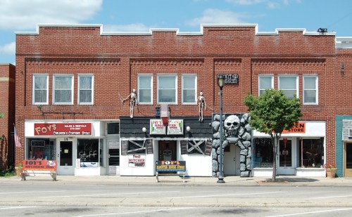 A row of brick storefronts, featuring a toy store, a Halloween shop, and a costume store, with decorative skeletons.