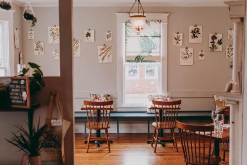 Cozy restaurant interior with wooden tables, chairs, and botanical art on the walls, featuring a bright window.