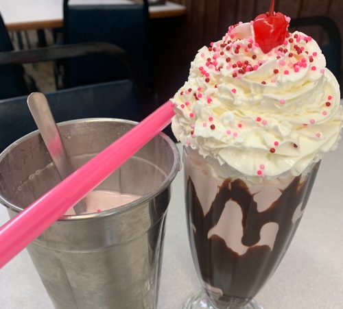 A tall glass of chocolate sundae topped with whipped cream, sprinkles, and a cherry, next to a metal cup with a pink straw.