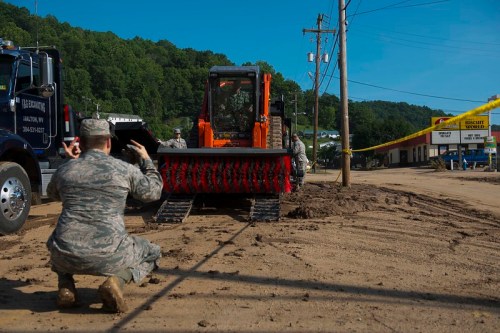 A soldier operates heavy machinery on a dirt road, with utility vehicles and power lines in the background.
