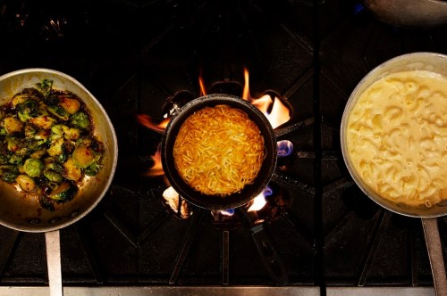 Three pots on a stove: sautéed Brussels sprouts, boiling noodles, and creamy macaroni and cheese, with flames visible.
