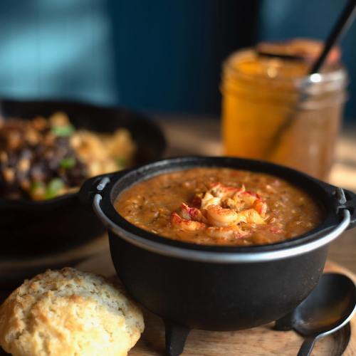 A bowl of shrimp gumbo with a biscuit, set on a wooden table with a drink in the background.