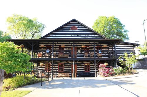 A two-story log cabin with a sloped roof, surrounded by greenery and colorful flowers, featuring a porch with rocking chairs.