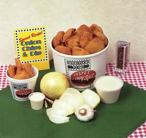 A display of onion chips with dip, an onion, garlic, and seasoning, on a checkered tablecloth.
