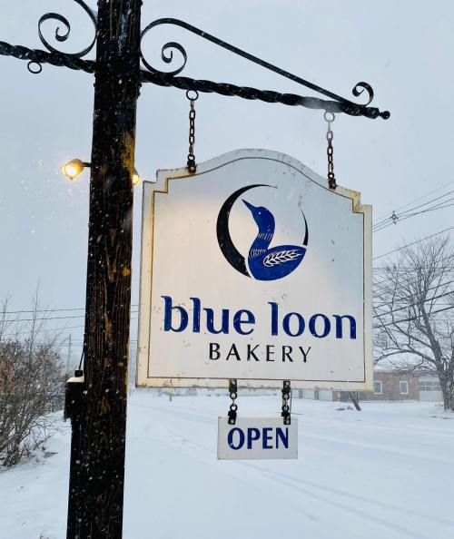 Sign for Blue Loon Bakery hanging outside, with "OPEN" displayed, amidst a snowy landscape.