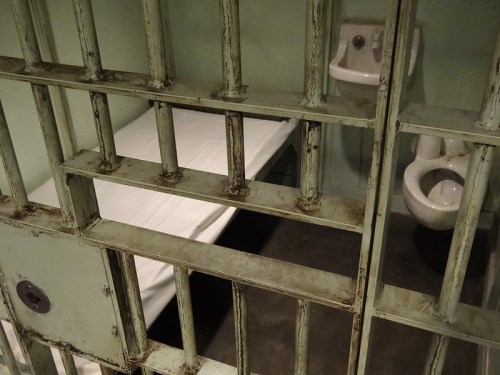 A view of a prison cell with a bed, toilet, and sink, seen through metal bars.