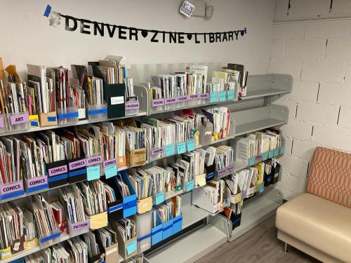 A shelf filled with various zines organized by category, with a sign reading "Denver Zine Library" above.