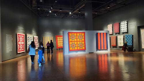 A gallery with colorful textile art on the walls and visitors exploring the exhibits.