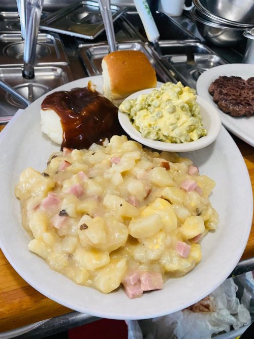 A plate of comfort food featuring creamy macaroni and cheese with ham, mashed potatoes, barbecue sauce, a roll, and salad.