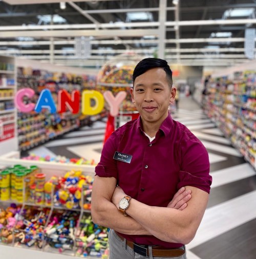 A smiling employee stands in a colorful candy aisle, with "CANDY" displayed prominently in the background.