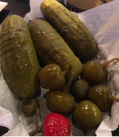 A basket of pickles, including large green pickles and small green and red peppers, on a bed of parchment paper.