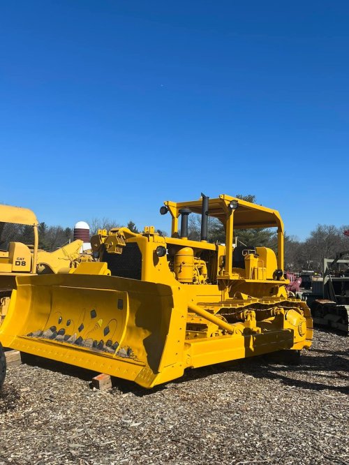 A bright yellow bulldozer parked on gravel under a clear blue sky, surrounded by construction equipment.