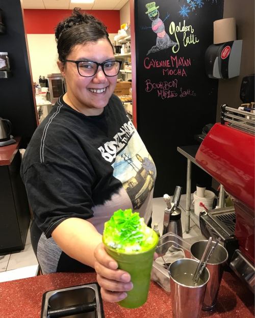 A smiling barista holds a green drink topped with whipped cream, standing behind a coffee counter.