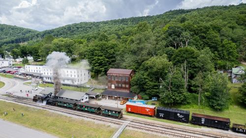 Aerial view of a vintage steam train at a station surrounded by lush green hills and a small town.