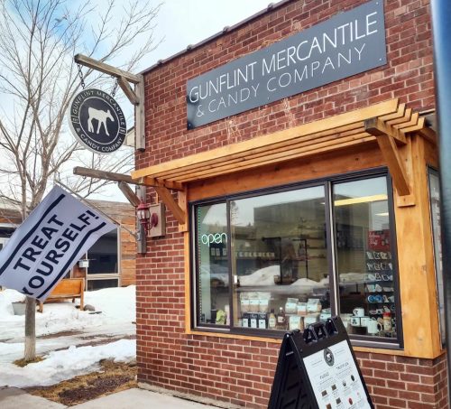 Exterior of Gunflint Mercantile & Candy Company, featuring a sign, open flag, and a menu board outside. Snow on the ground.