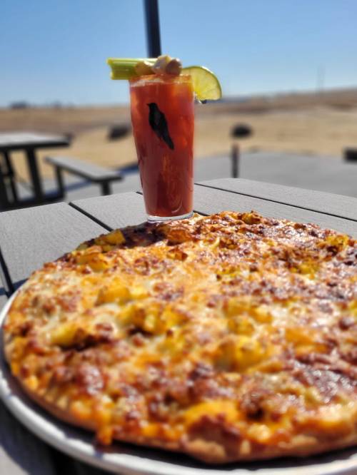 A close-up of a cheesy pizza on a table, with a cocktail garnished with celery and lime in the background.