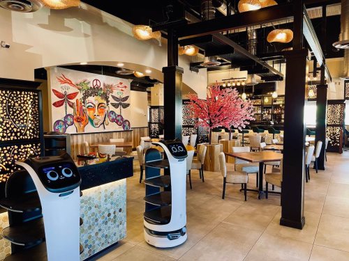 A modern restaurant interior featuring robotic servers, elegant decor, and a vibrant mural of a face with floral elements.