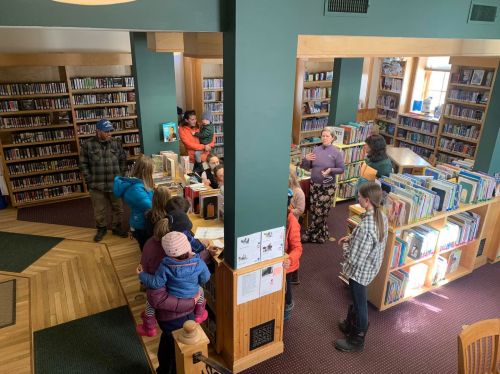 A busy library scene with people gathered around a central desk, surrounded by bookshelves filled with books.