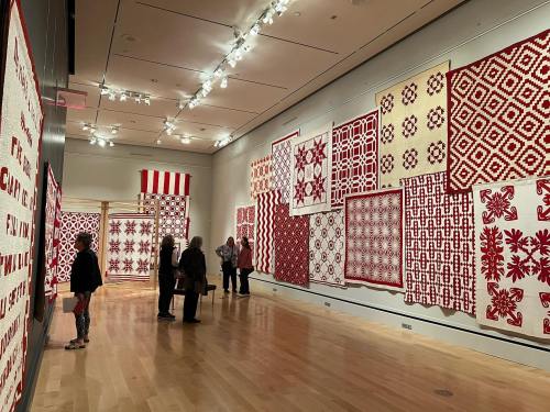 Visitors admire a gallery wall adorned with various red and white quilts in a spacious art museum.