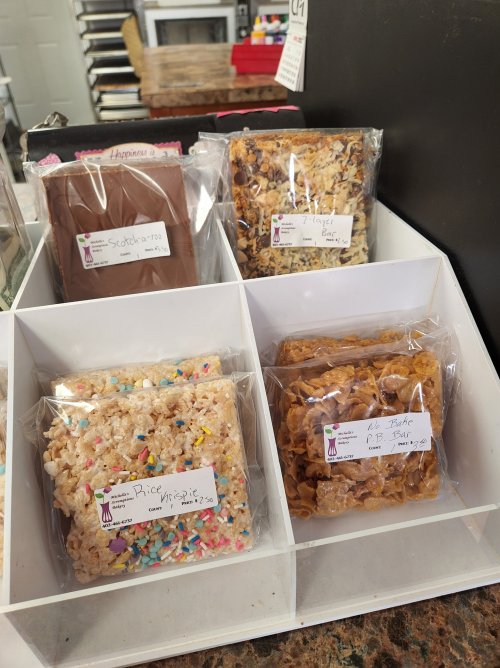 A display of assorted treats in clear packaging, including Rice Krispie treats and layered bars, on a counter.