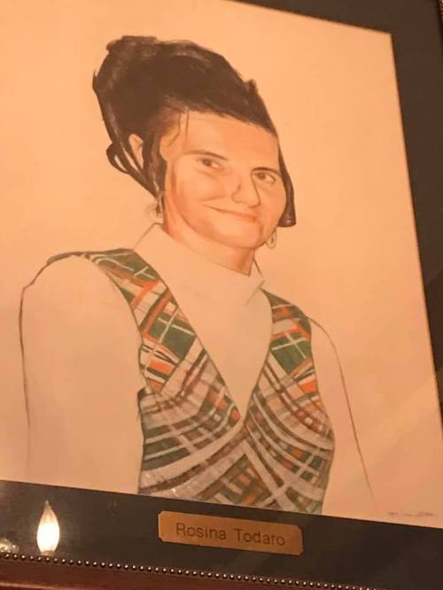 A framed portrait of a woman with dark hair, wearing a patterned vest and a white shirt, labeled "Rosina Todaro."