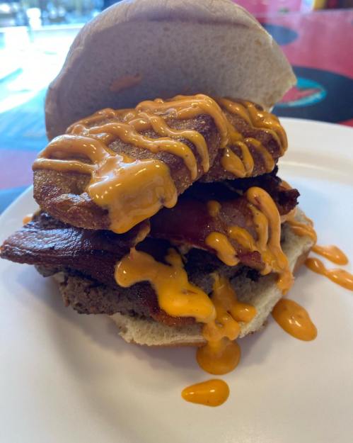 A close-up of a sandwich stacked with crispy bacon, fried chicken, and drizzled with a creamy sauce on a bun.