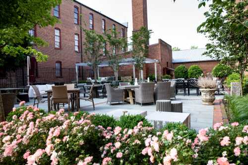 Outdoor dining area with tables and chairs surrounded by greenery and flowers, set against a brick building backdrop.