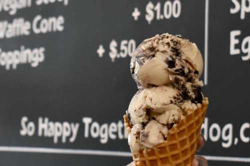 A hand holds a waffle cone filled with two scoops of cookies and cream ice cream against a menu backdrop.