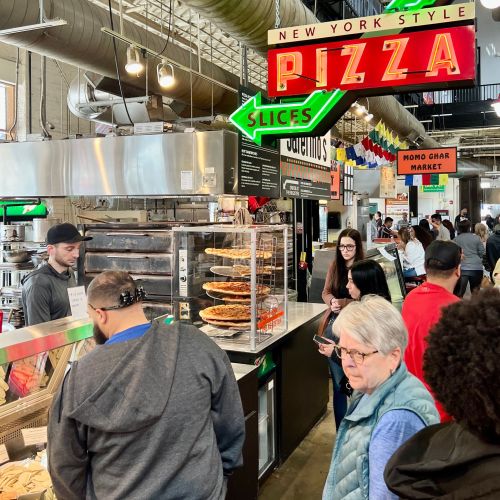 A busy food market with people ordering pizza and other items, featuring a bright "New York Style Pizza" sign.