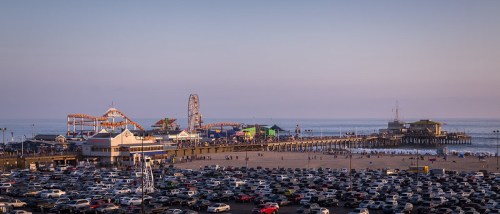 A vibrant pier with amusement rides at sunset, overlooking a crowded beach and a parking lot filled with cars.