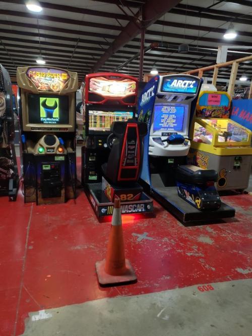 A row of arcade machines, including racing and sports games, with a traffic cone in front on a red floor.