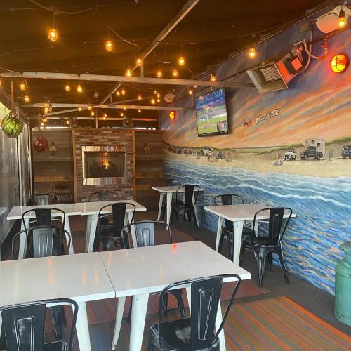 Cozy restaurant interior with a beach mural, string lights, a fireplace, and empty tables. A TV displays a sports game.