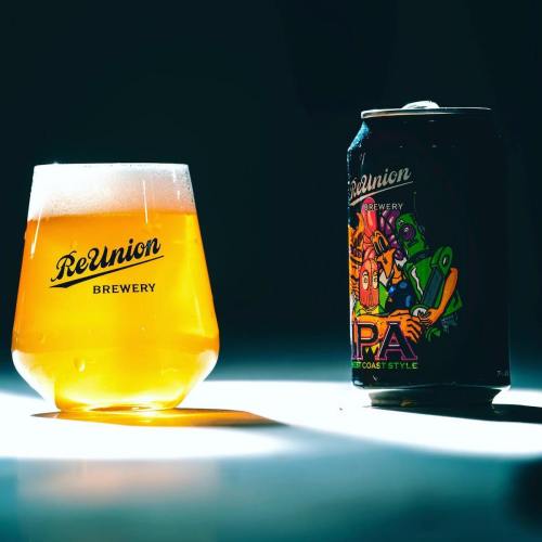 A glass of golden beer next to a colorful IPA can, both featuring the ReUnion Brewery logo.