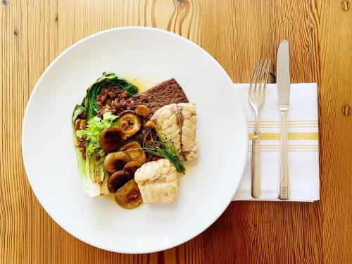 A plate of fish with mushrooms, greens, and a slice of bread, set on a wooden table with cutlery beside it.