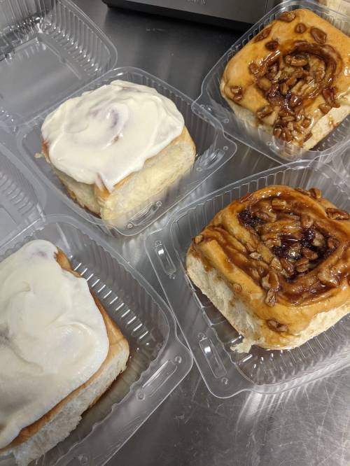 Four delicious cinnamon rolls in clear containers, topped with cream cheese frosting and caramelized nuts.