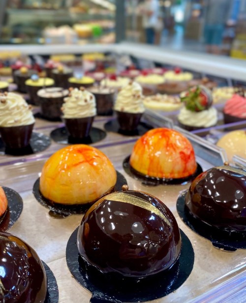 A variety of colorful desserts, including glossy chocolate and fruit-flavored dome cakes, displayed in a pastry case.