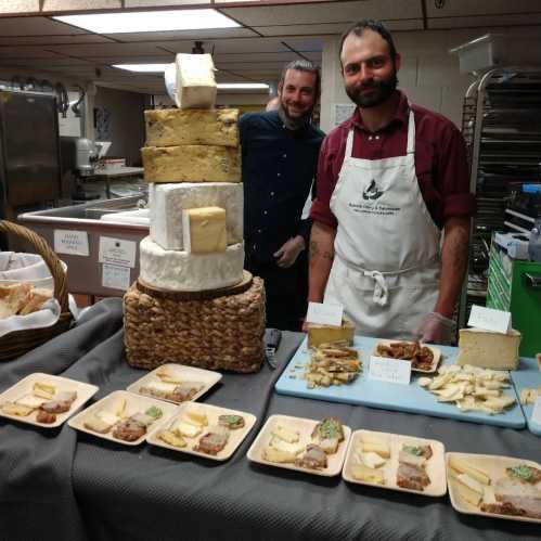 Two chefs stand behind a display of cheese, featuring a tall cheese tower and various cheese samples on plates.