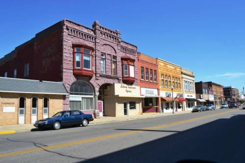 Historic buildings line a sunny street, showcasing a mix of architectural styles and storefronts in a small town.