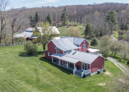 Aerial view of a red farmhouse with a metal roof, surrounded by green fields and trees in a rural setting.