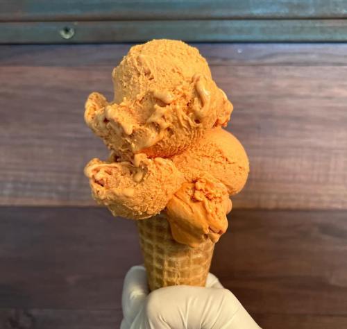 A hand holding a waffle cone topped with two scoops of orange ice cream against a wooden background.
