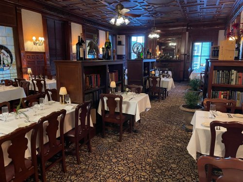Elegant restaurant interior with white tablecloths, wooden furniture, and decorative bookshelves. Warm, inviting atmosphere.